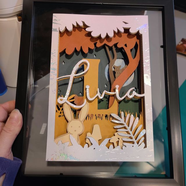 Cadre de naissance en carton 3D : prénom « Livia » en lettres argentées devant un grand L, avec lapin, arbre et feuillages en couches superposées.