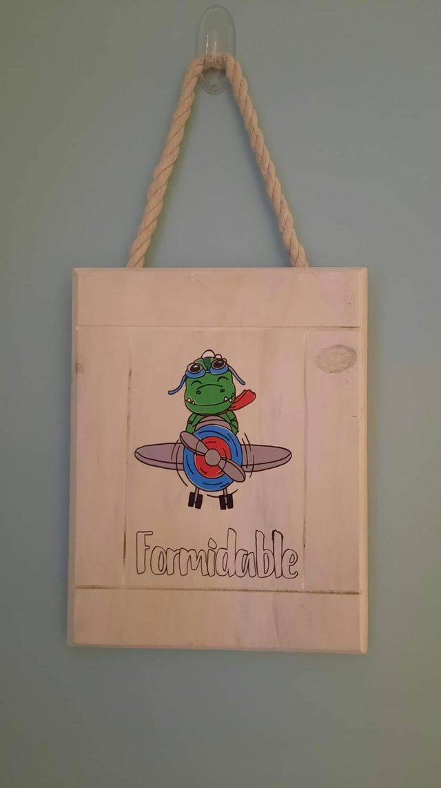 Plaque en bois blanchi suspendue par une corde : petit dinosaure vert aux commandes d'un avion, avec le mot « Formidable ».