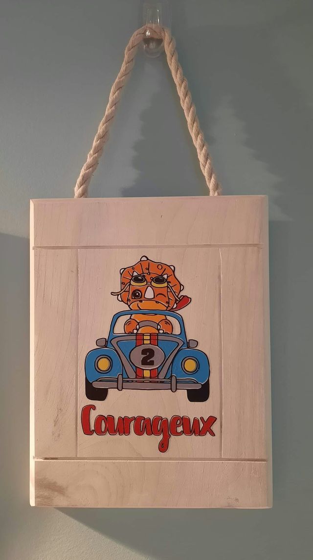 Plaque en bois blanchi suspendue par une corde : tricératops conduisant une voiture de course bleue, avec le mot « Courageux » en rouge.