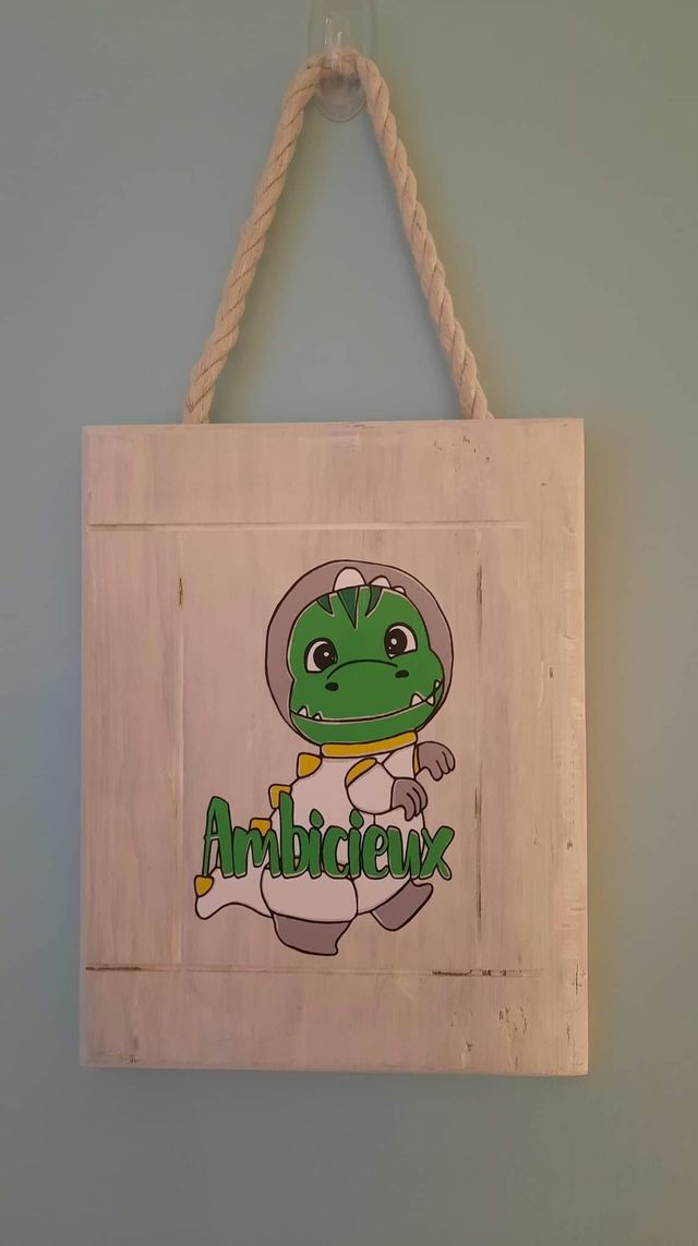 Plaque en bois blanchi suspendue par une corde : tyrannosaure vert en costume d'astronaute, avec un mot en lettres vertes.