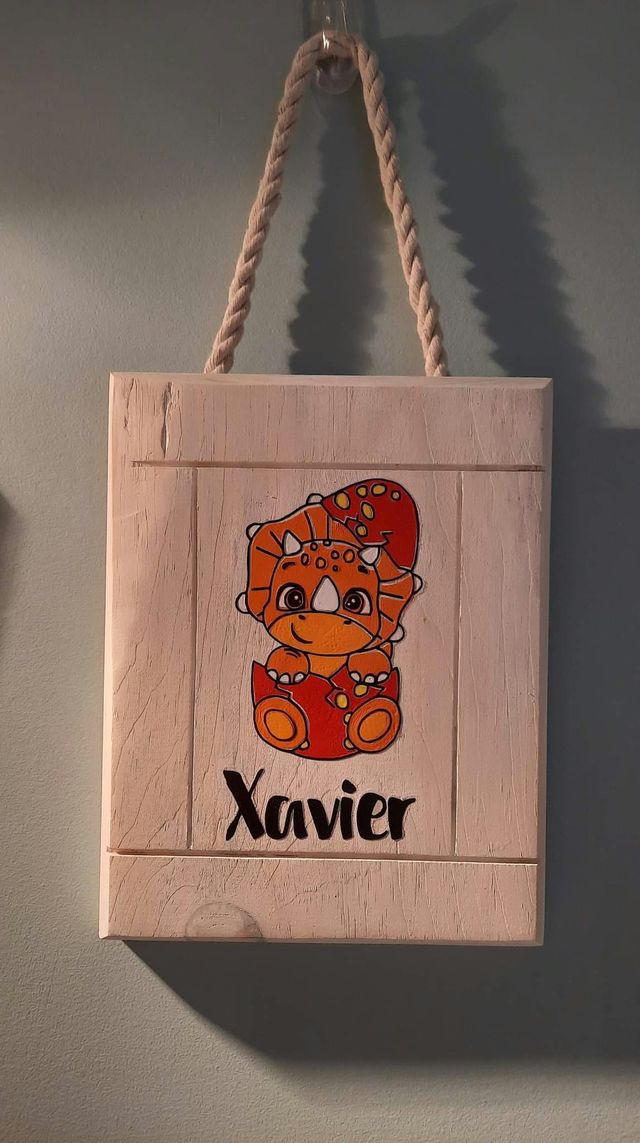 Plaque en bois blanchi suspendue par une corde : bébé tricératops orange sortant de son œuf, avec le prénom « Xavier ».