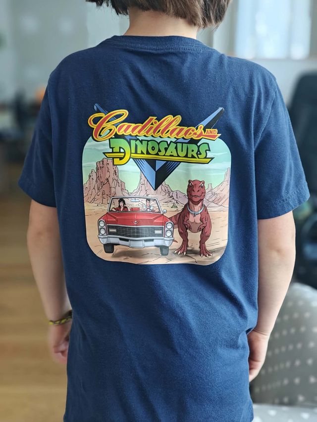 Dos d'un t-shirt bleu marine porté par un enfant, orné d'une grande illustration colorée « Cadillacs and Dinosaurs » : une décapotable rouge et un tyrannosaure dans le désert.