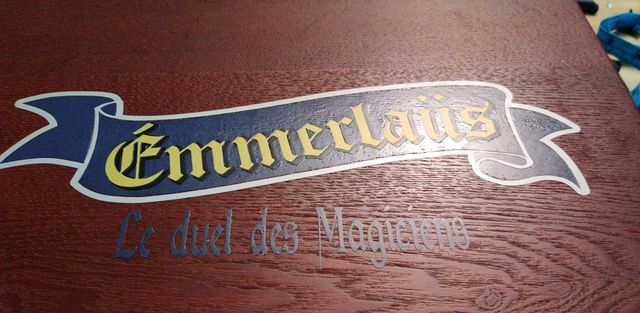 Couvercle de boîte en bois foncé décoré d'une banderole en vinyle « Emmerlaüs, le duel des magiciens ».