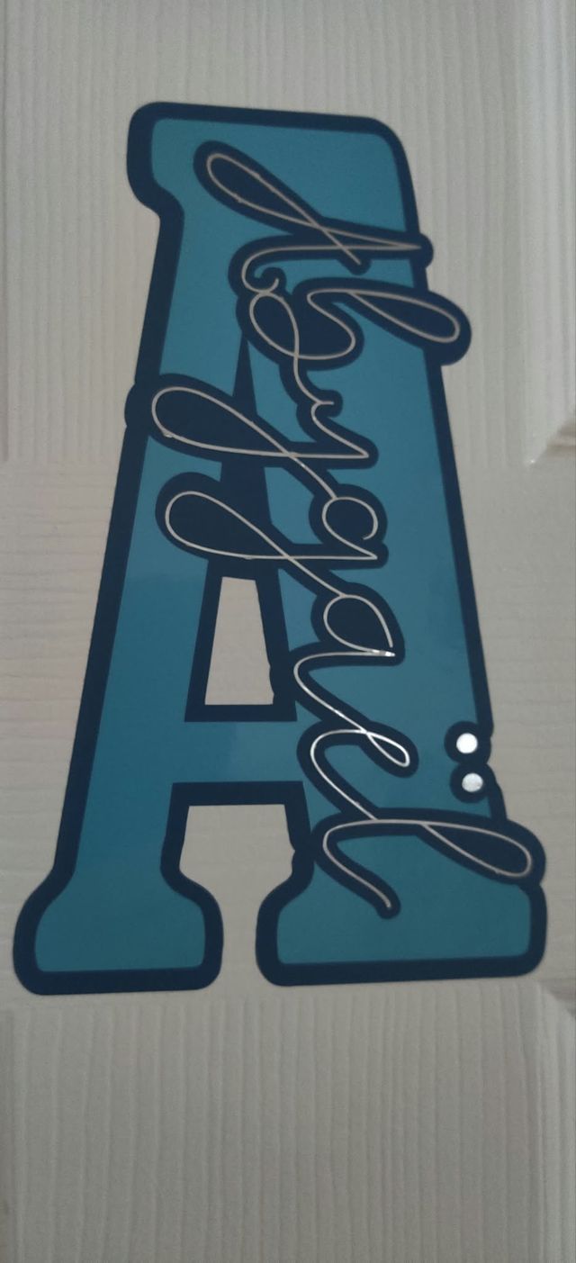 Grande lettre A turquoise découpée, accrochée à une porte, avec le prénom « Abygaël » en lettres argentées.