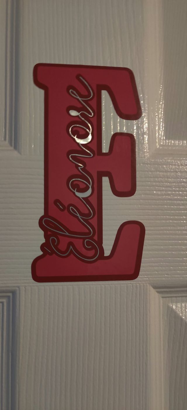 Grande lettre E rouge découpée, accrochée à une porte, avec le prénom « Éléonore » en lettres argentées.
