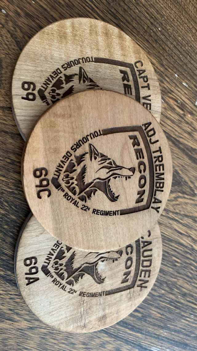 Trois sous-verres ronds en bois gravés d'une tête de loup et de l'inscription « Toujours devant, Recon, Royal 22e Régiment », chacun au nom d'un militaire.